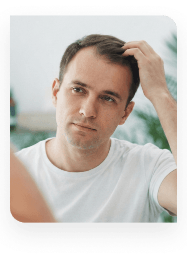 A front view of a man in a white t-shirt looking at his reflection in a mirror while running his fingers through his hair, inspecting his hairline or scalp concern.